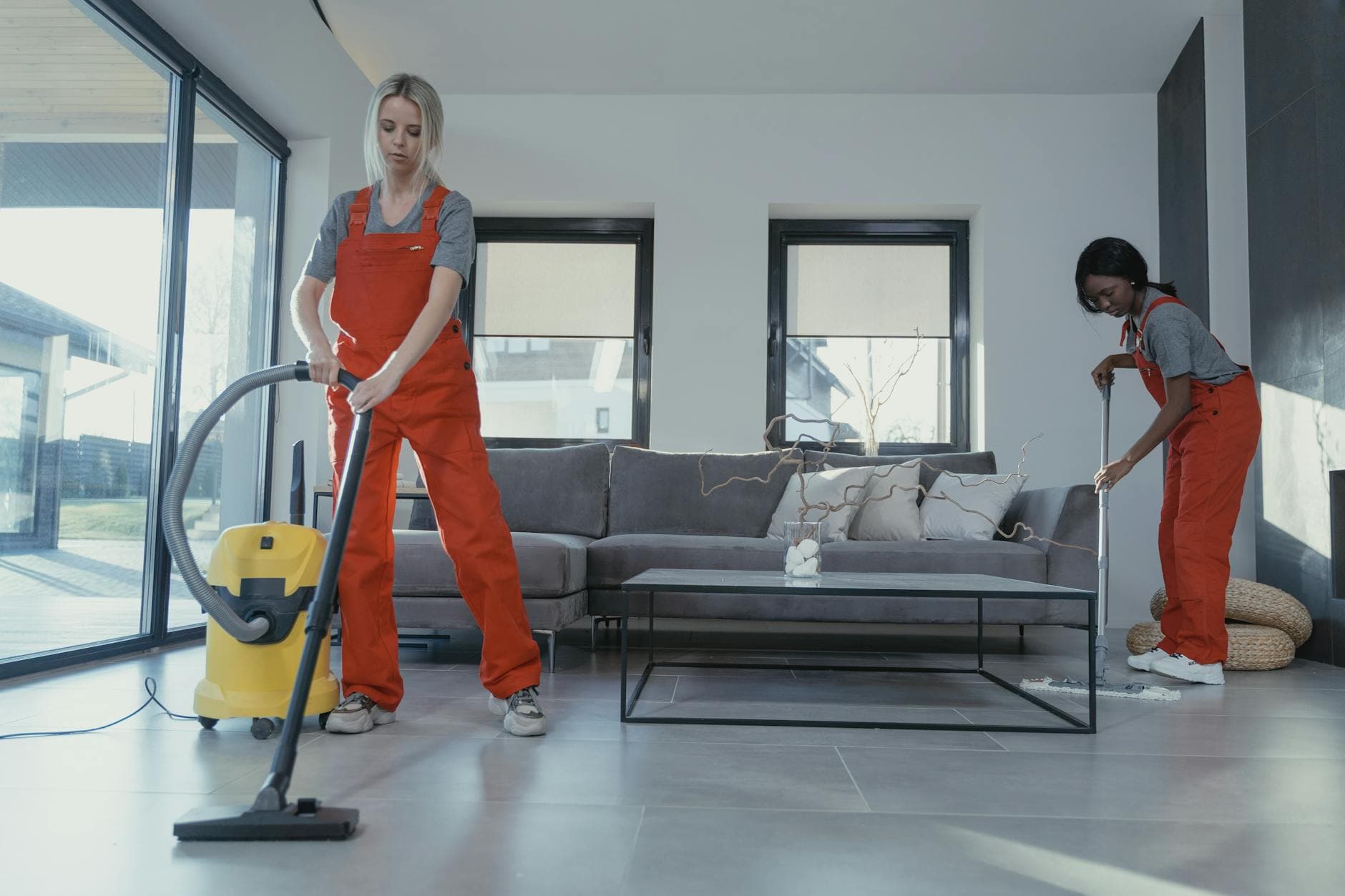 Two NYC Maid cleaners in red overalls vacuuming and sweeping a modern NYC living room — Post-Construction Cleanup in Stapleton, Staten Island